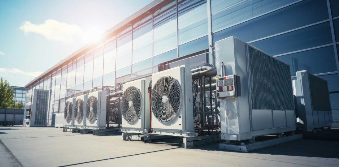 Reihe industrieller Verflüssiger mit Ventilatoren und Rohrleitungen vor der Glasfassade eines Gewerbegebäudes.