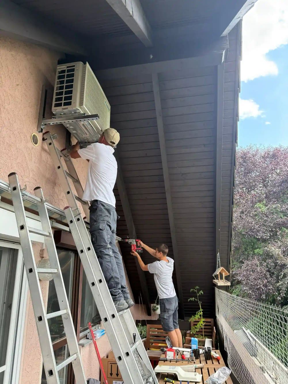 Zwei Monteure setzen ein Außengerät an einer Hauswand, einer auf der Leiter, einer am Boden.